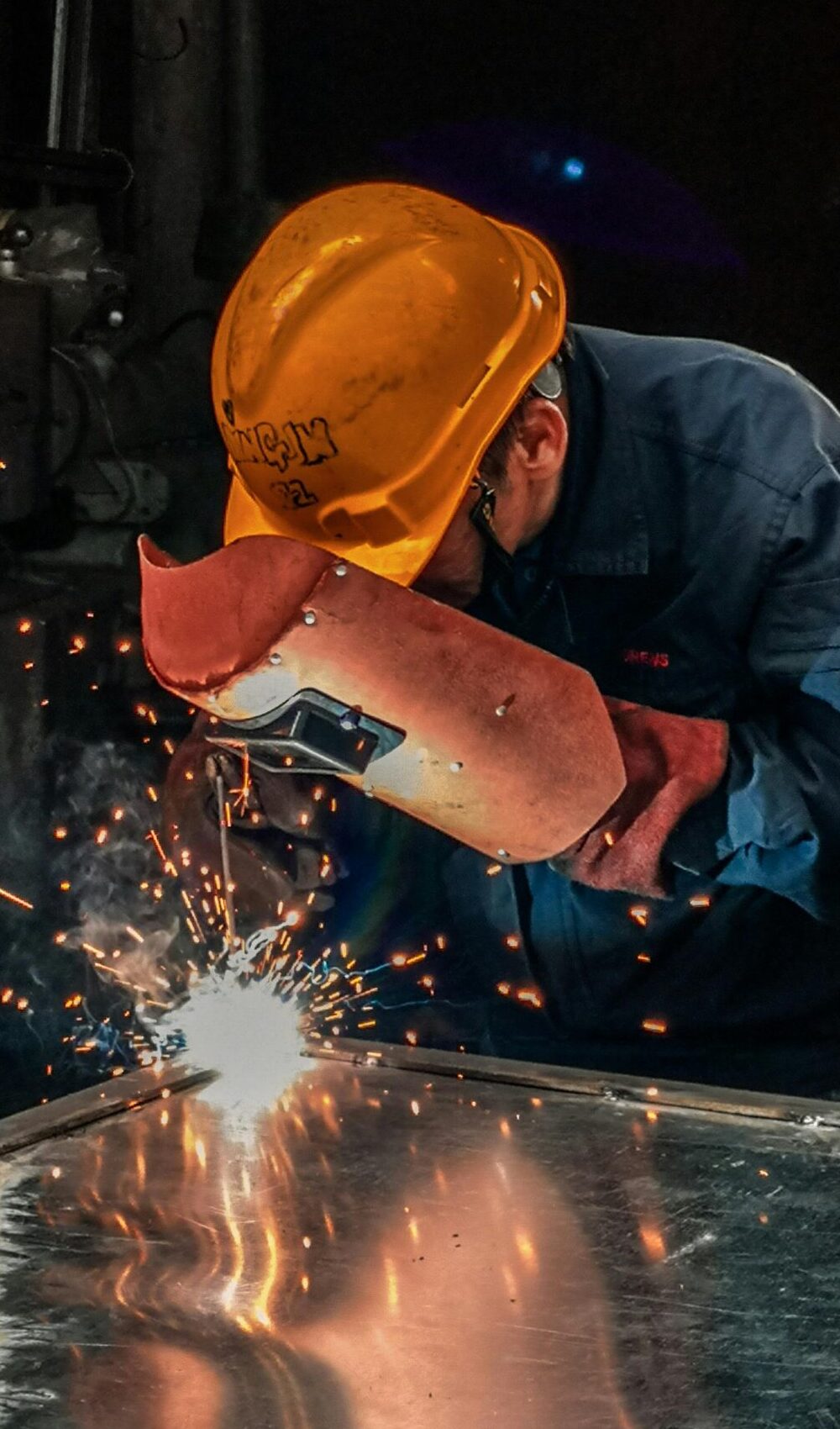 A welder wearing protective gear works on metal in an industrial workshop, surrounded by sparks.